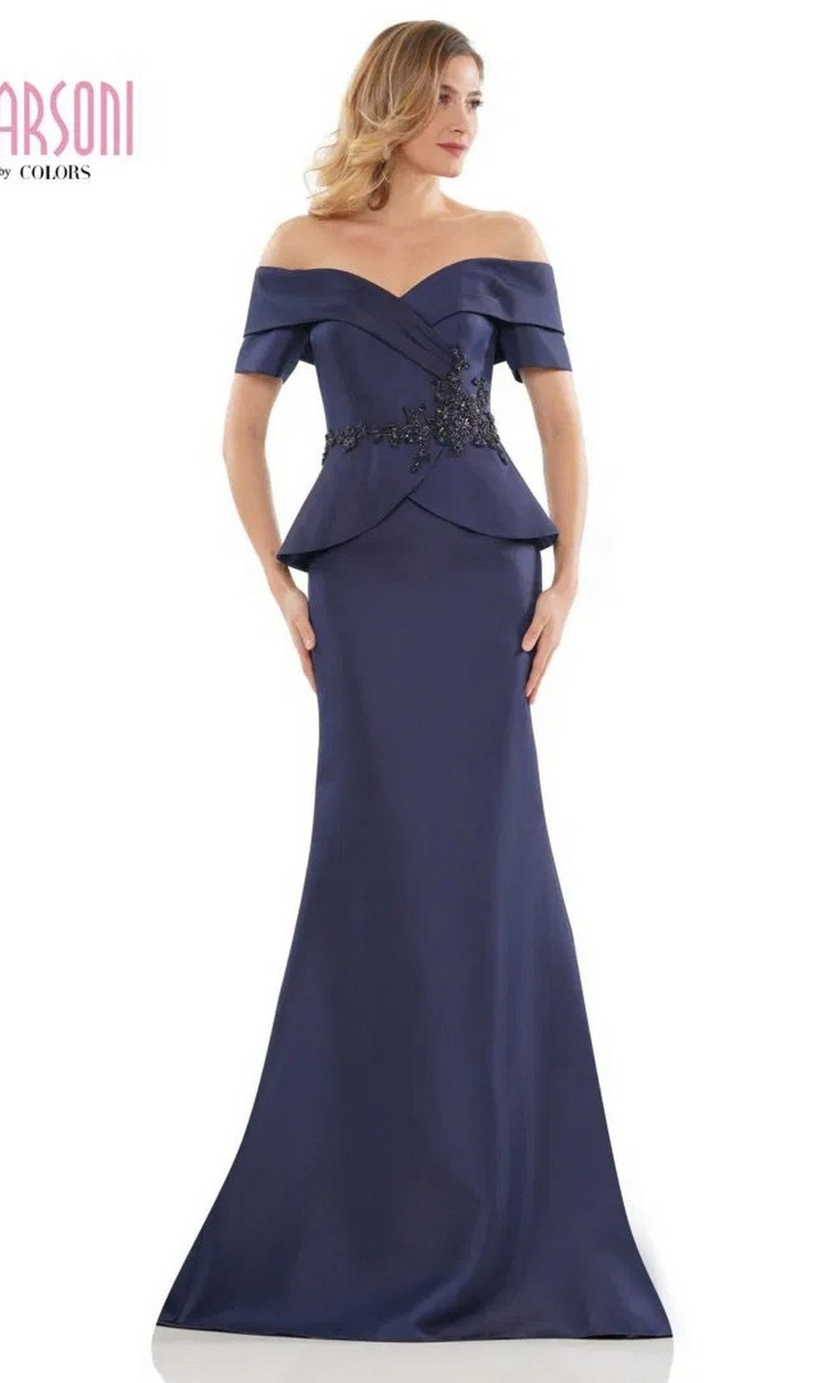 A woman wears the Marsoni MV1144 Long Formal Dress in navy, featuring an off-the-shoulder neckline, fitted bodice, and black floral waist embellishments for an elegant floor-length silhouette.